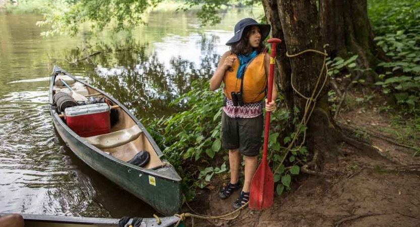 A young person stands on the shore of a river holding a paddle. There is a canoe floating nearby. 